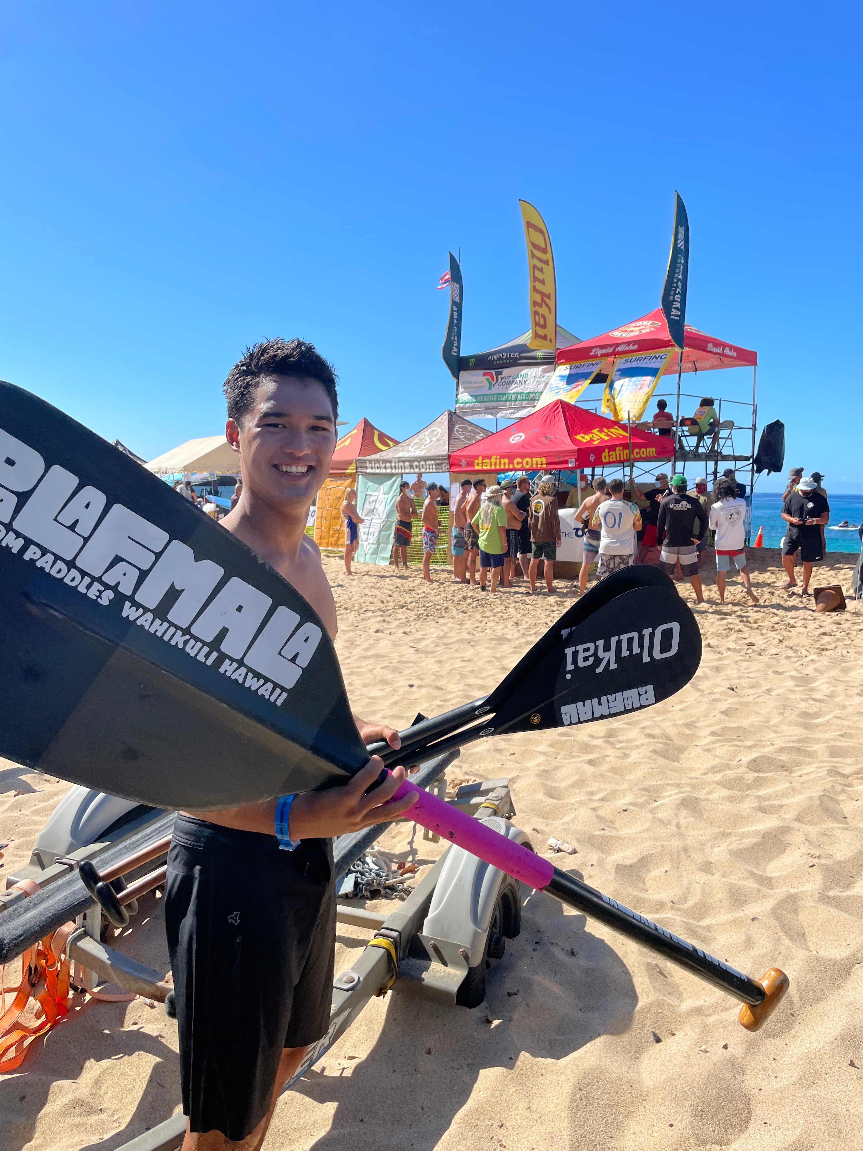 Person holding a paddle with Olukai branding on a beach with spectators and tents in the background.